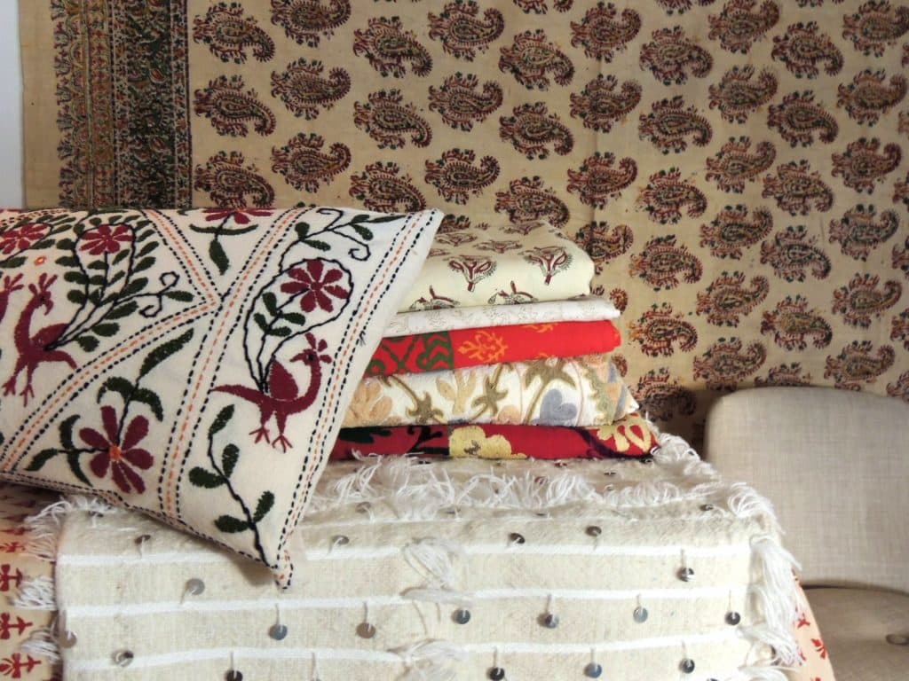 A vintage hand-blocked red and brown Kalamkari cotton paisley coverlet/cloth hangs in the background. On the table is a stack of vintage hand-blocked Indian bed covers and vintage embroidered suzanis from Uzbekistan, and a pillow made from vintage Indian colorful floral embroidered fabric, laying on top of a Moroccan beige and white wedding blanket with white fringe and silver sequins. All available from Antique Textiles Galleries.