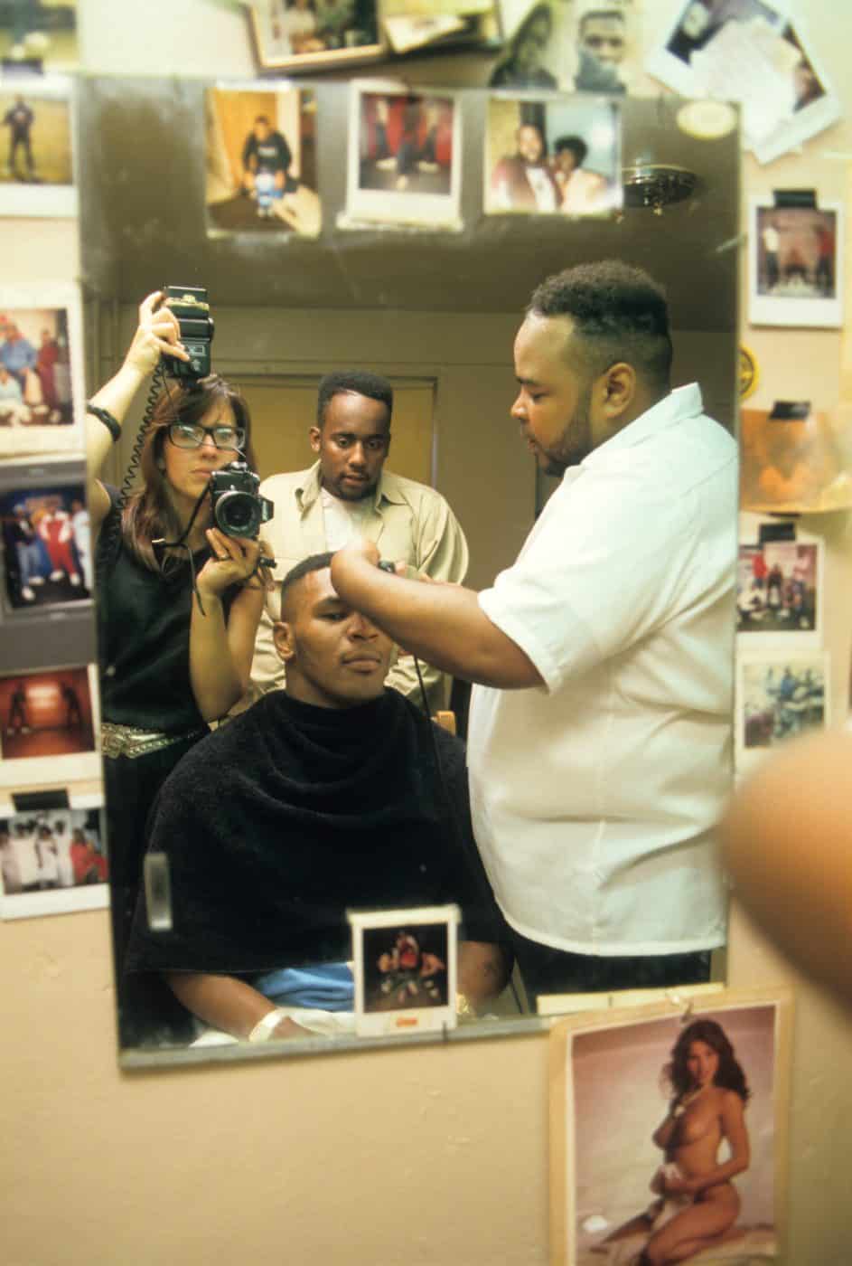 Lori Grinker (reflected in the mirror) photographs Mike Tyson getting his hair cut in his boyhood neighborhood of Brownsville, Brooklyn, 1987
