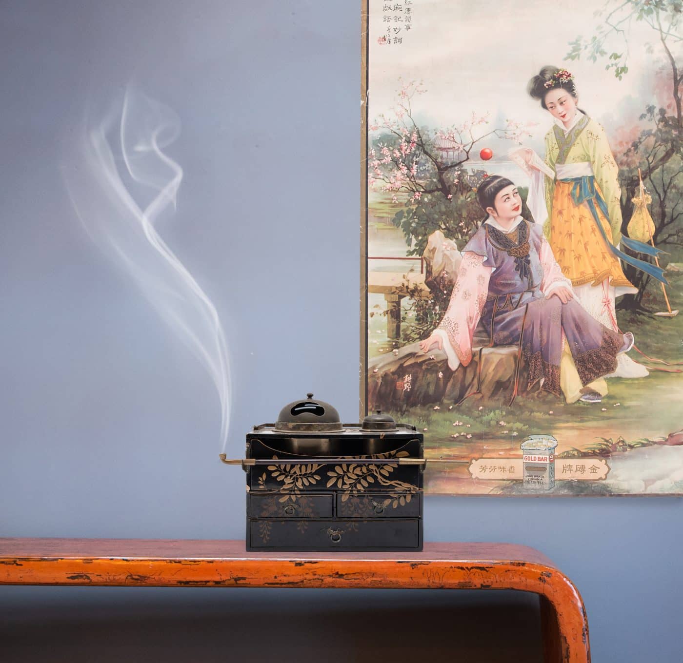 A mid-19th-century Japanese TAbako-bon, or tobacco tray, is decorated with lacquer and gilt in the takamaki-e style. Behind it is a 1930s Shanghai Art Deco poster advertising Gold Bar Cigarettes.