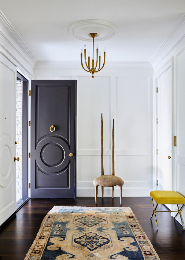 A white-walled entryway designed by Jeff Schlarb with a sculptural chair, a yellow velvet stool and a Persian rug