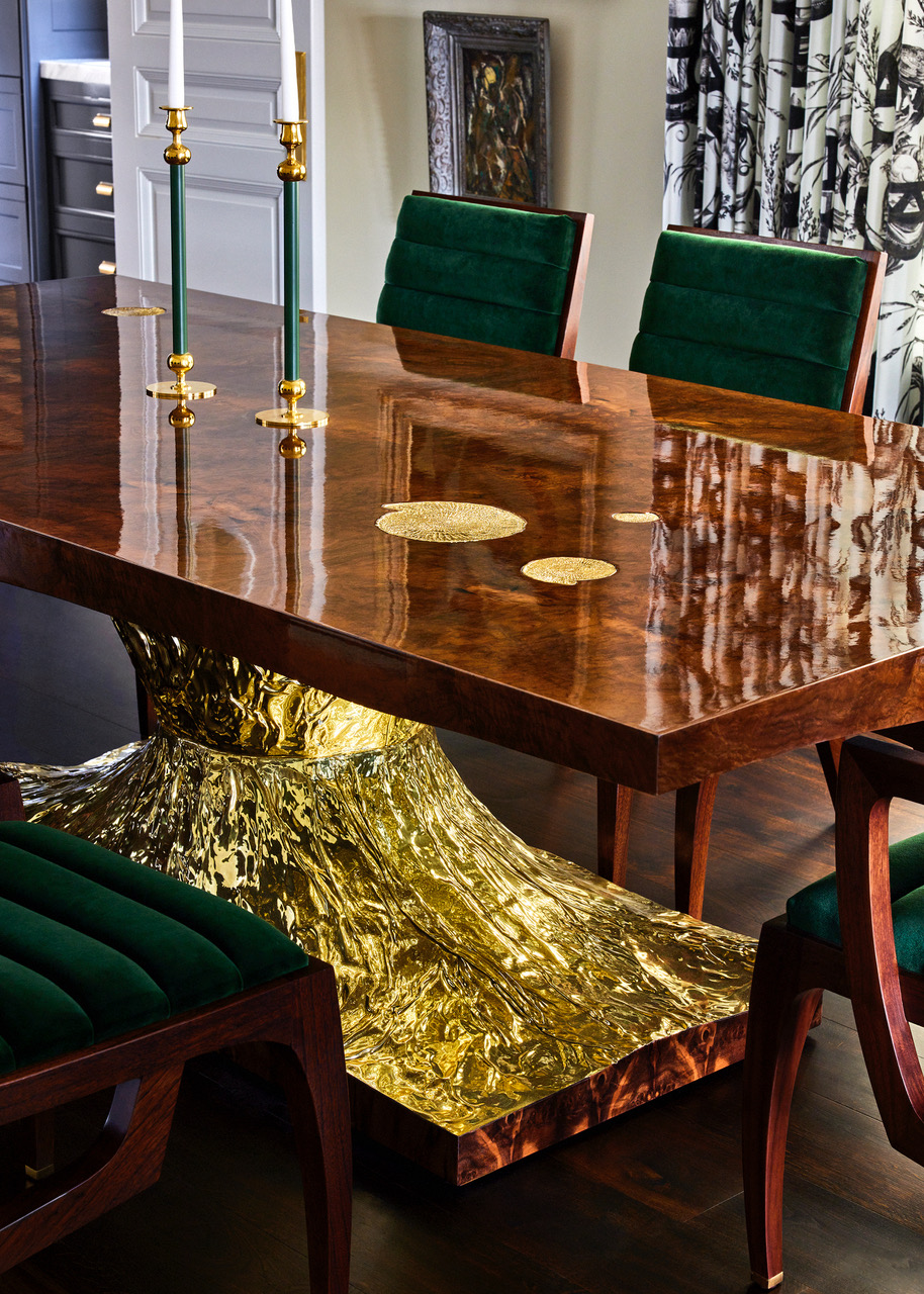 A dining room designed by Jeff Schlarb with a glossy, walnut-veneered dining table with a sculpted brass base surrounded by green velvet dining chairs