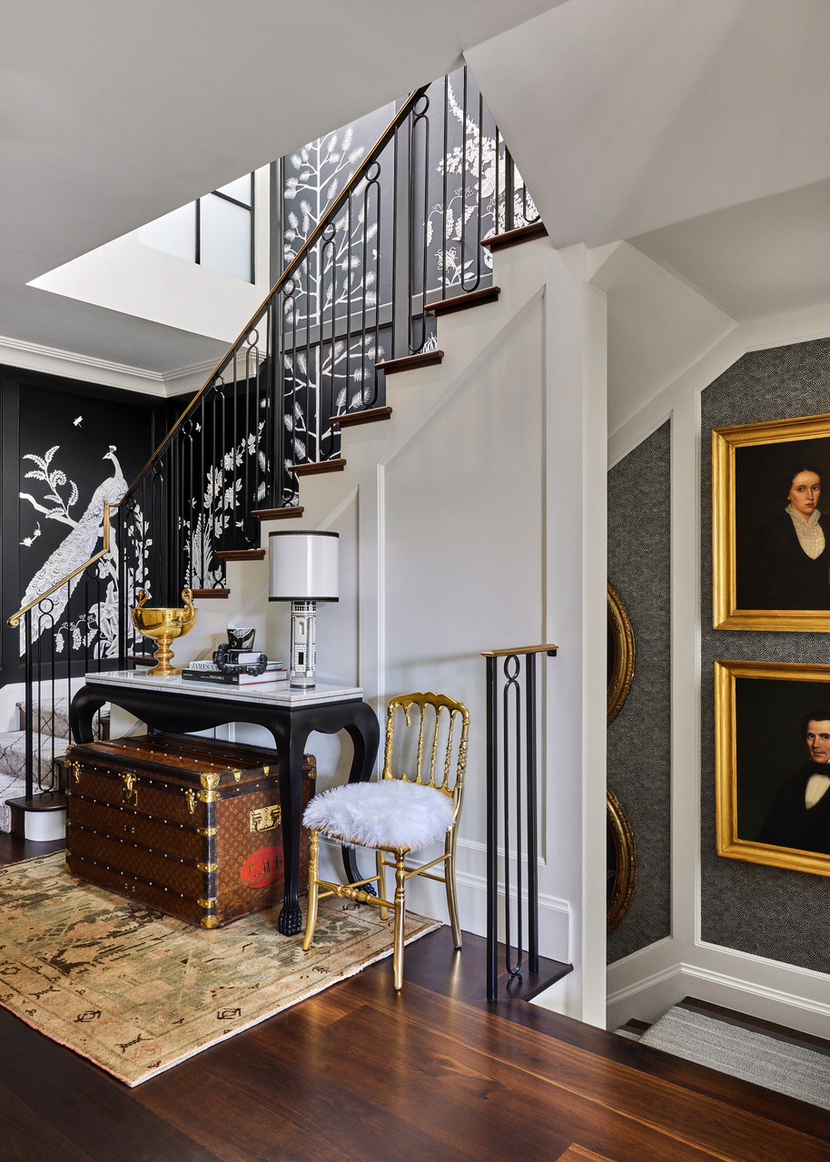 A stairway landing designed by Jeff Schlarb with a Louis Vuitton trunk under a marble-topped console table, next to a gilded chair with a seat upholstered in a fluffy white fabric