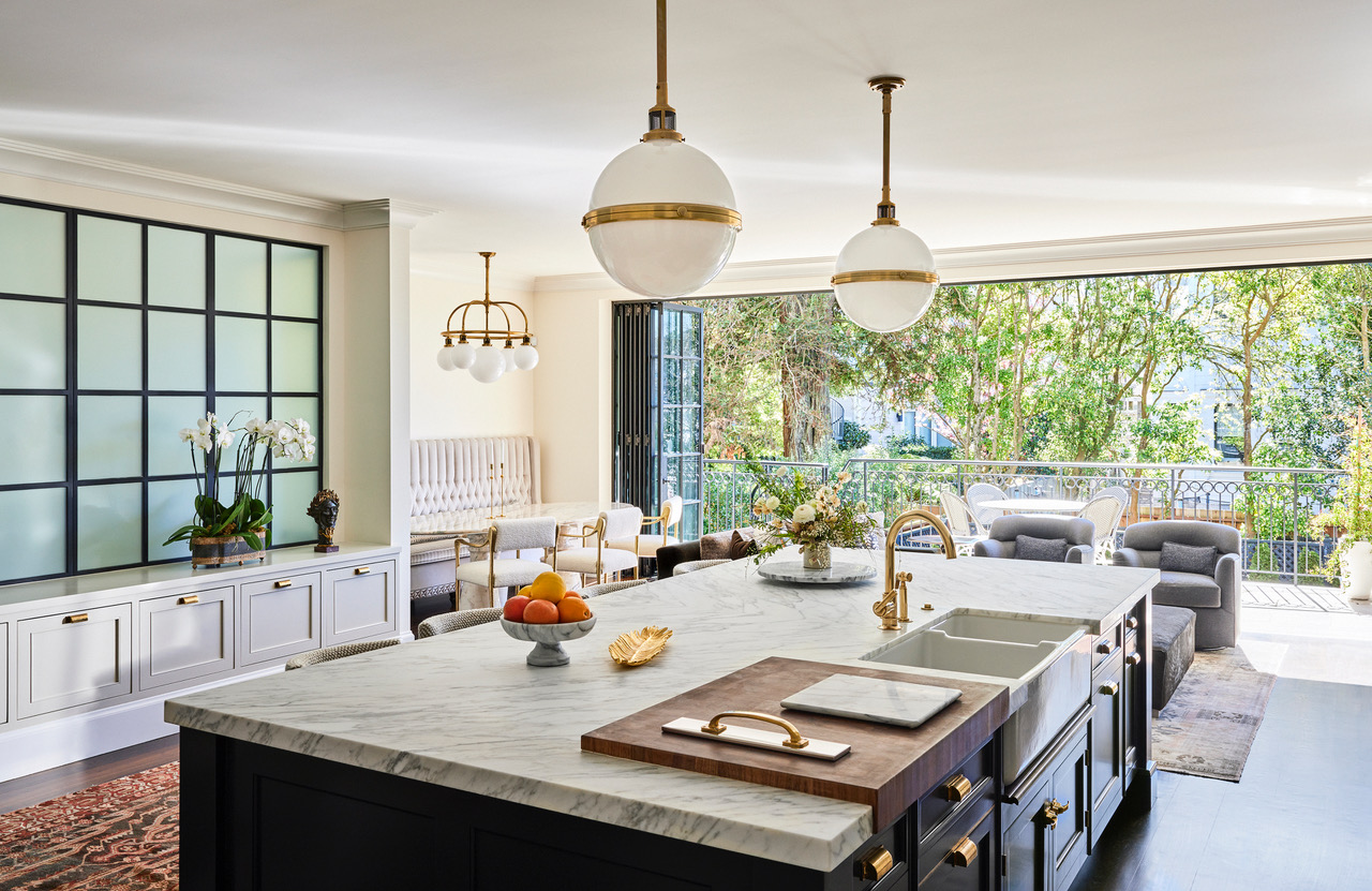 A large kitchen designed by Jeff Schlarb with a marble-topped kitchen island, a seating area with two gray lounge chairs, a breakfast nook with a white banquette and oval dining table, and folding doors opening onto a wide terrace