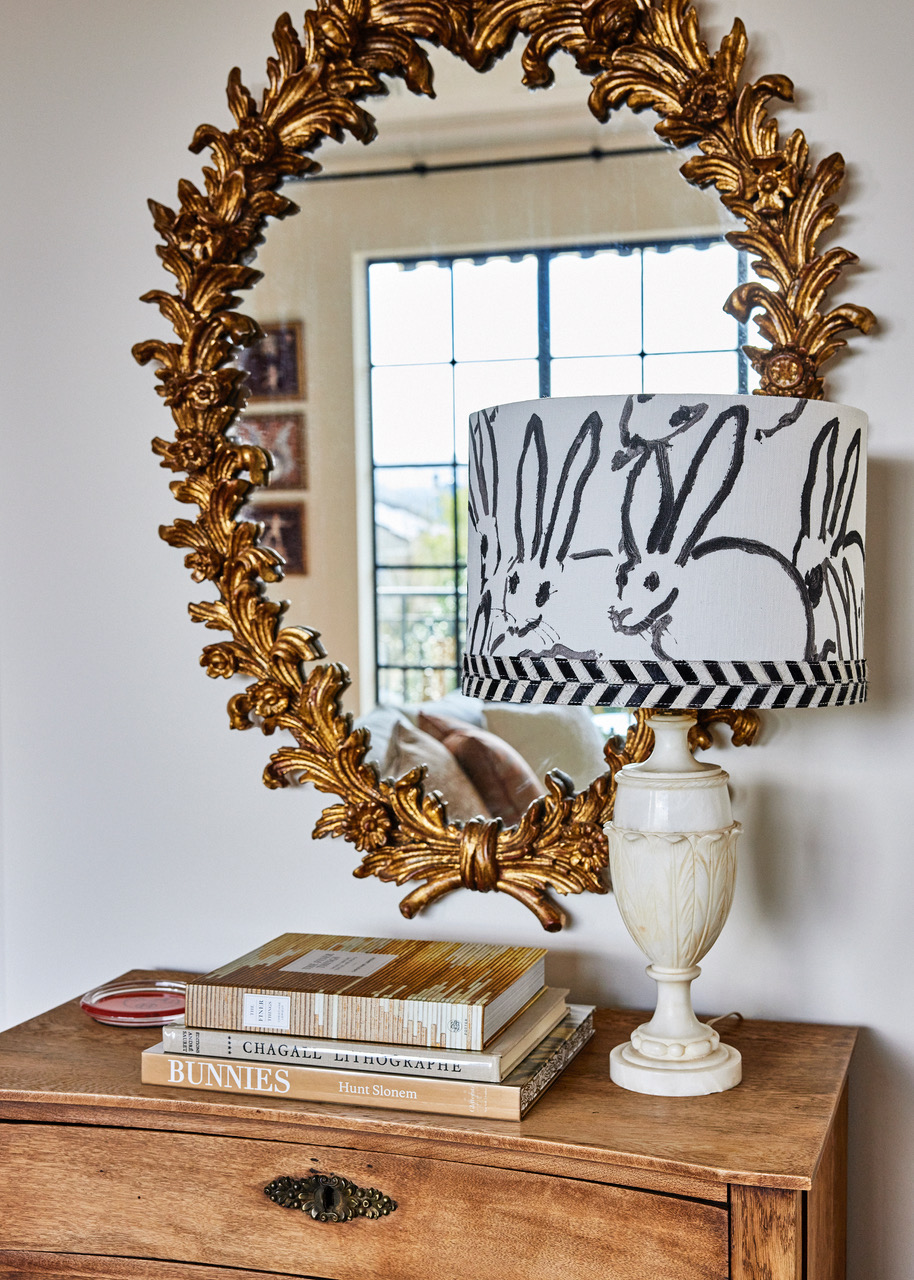 A gilded wall mirror and a lamp with a shade decorated with Hunt Slonem bunnies in a room designed by Jeff Schlarb