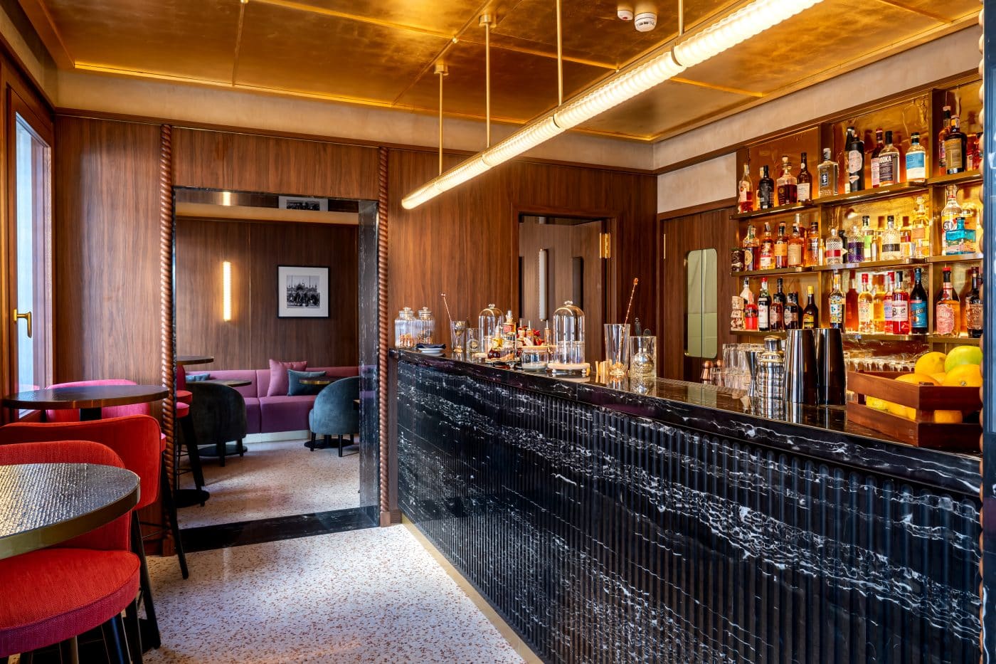 Venice's new Ca' di Dio hotel bar with terrazzo floor, black marble bar front and red upholstered chairs at round tables. Hotel architecture and interior design by Patricia Urquiola