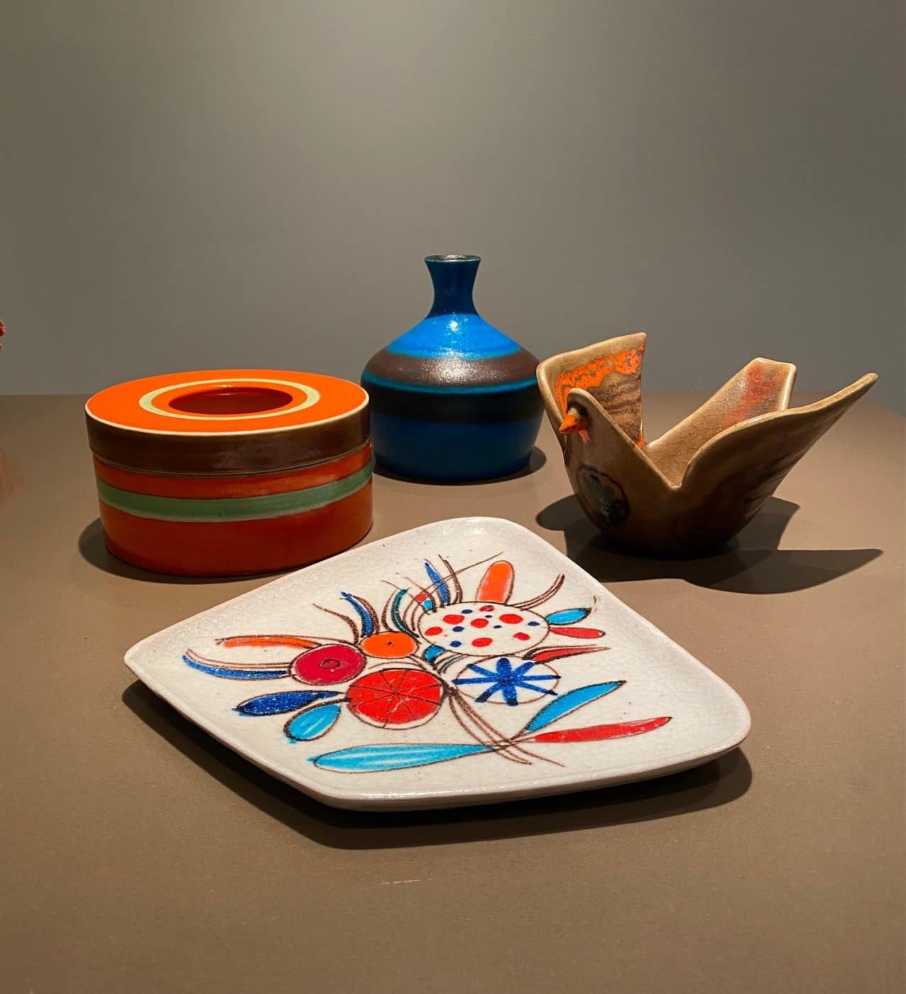 A white plate with abstract red orange and blue flowers by Guido Gambone surrounded by other ceramic vessels