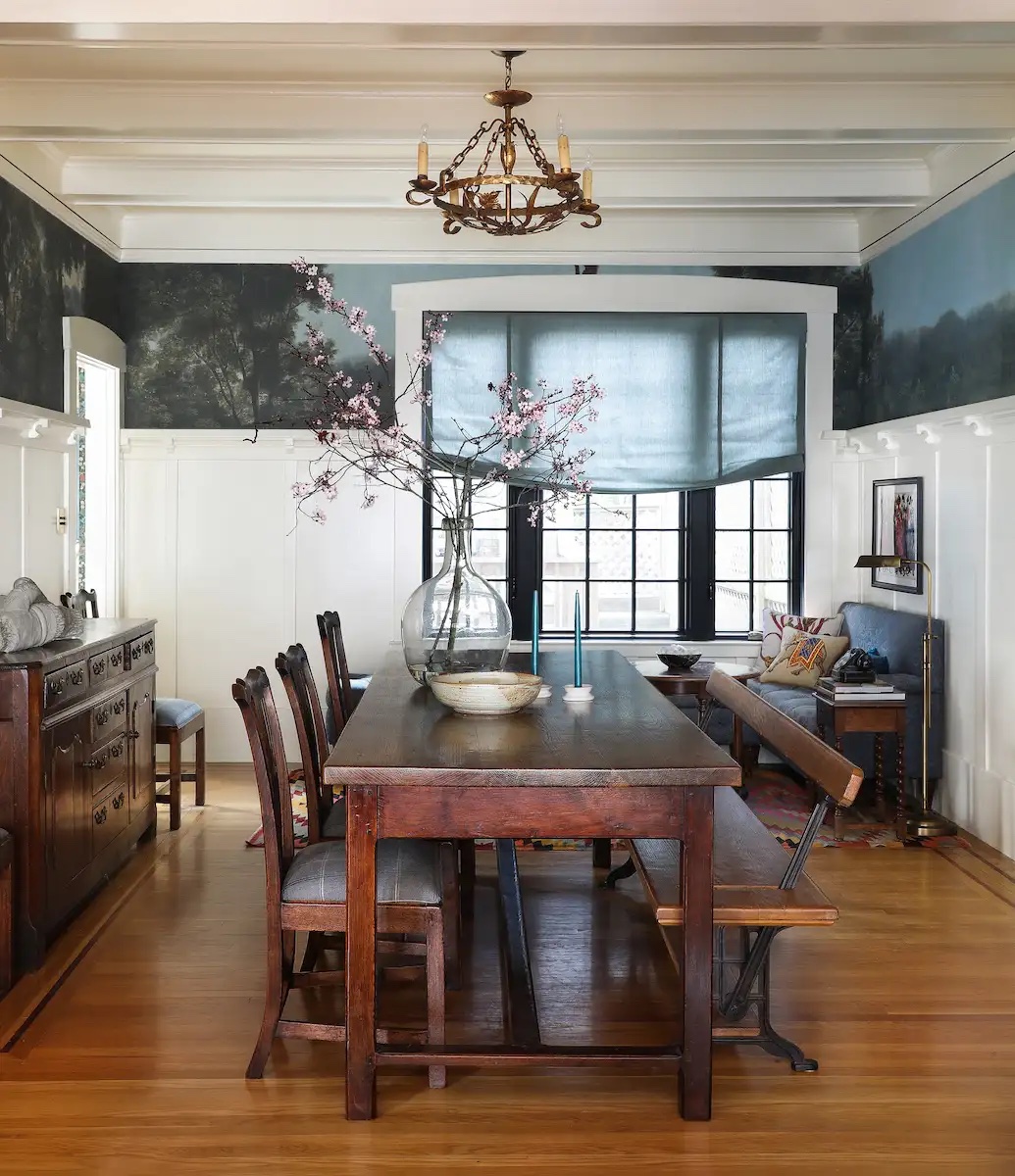 A dining room with dark wood furniture, warm-white walls and a frieze mural of green trees and a blue sky