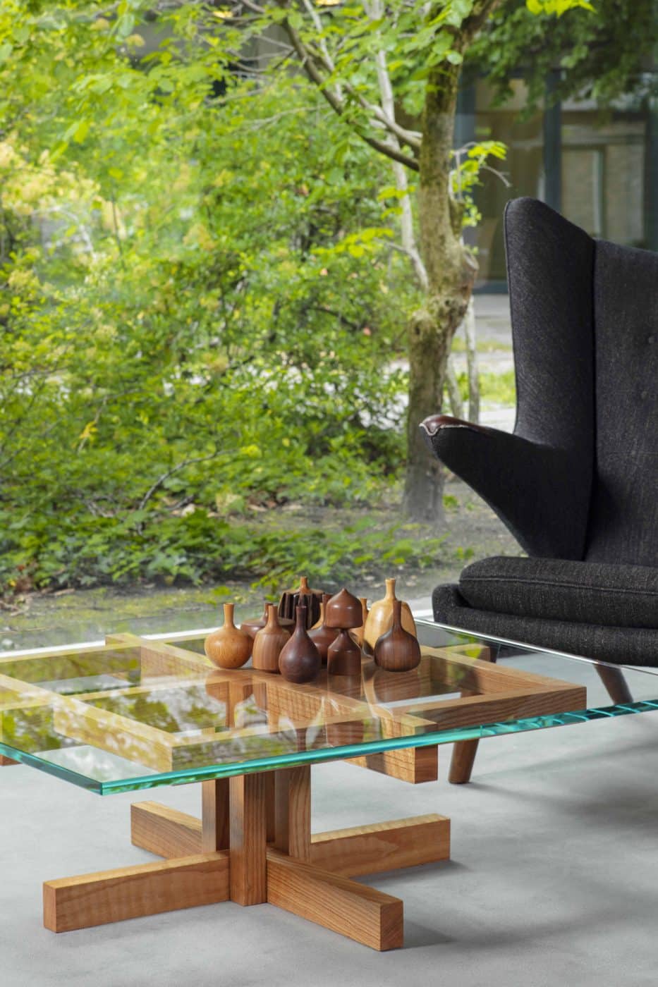 A glass-topped Ray Kappe coffee table displaying a collection of small wooden vases sits next to a dark-gray armchair and a floor-to-ceiling window with a view of green trees and bushes