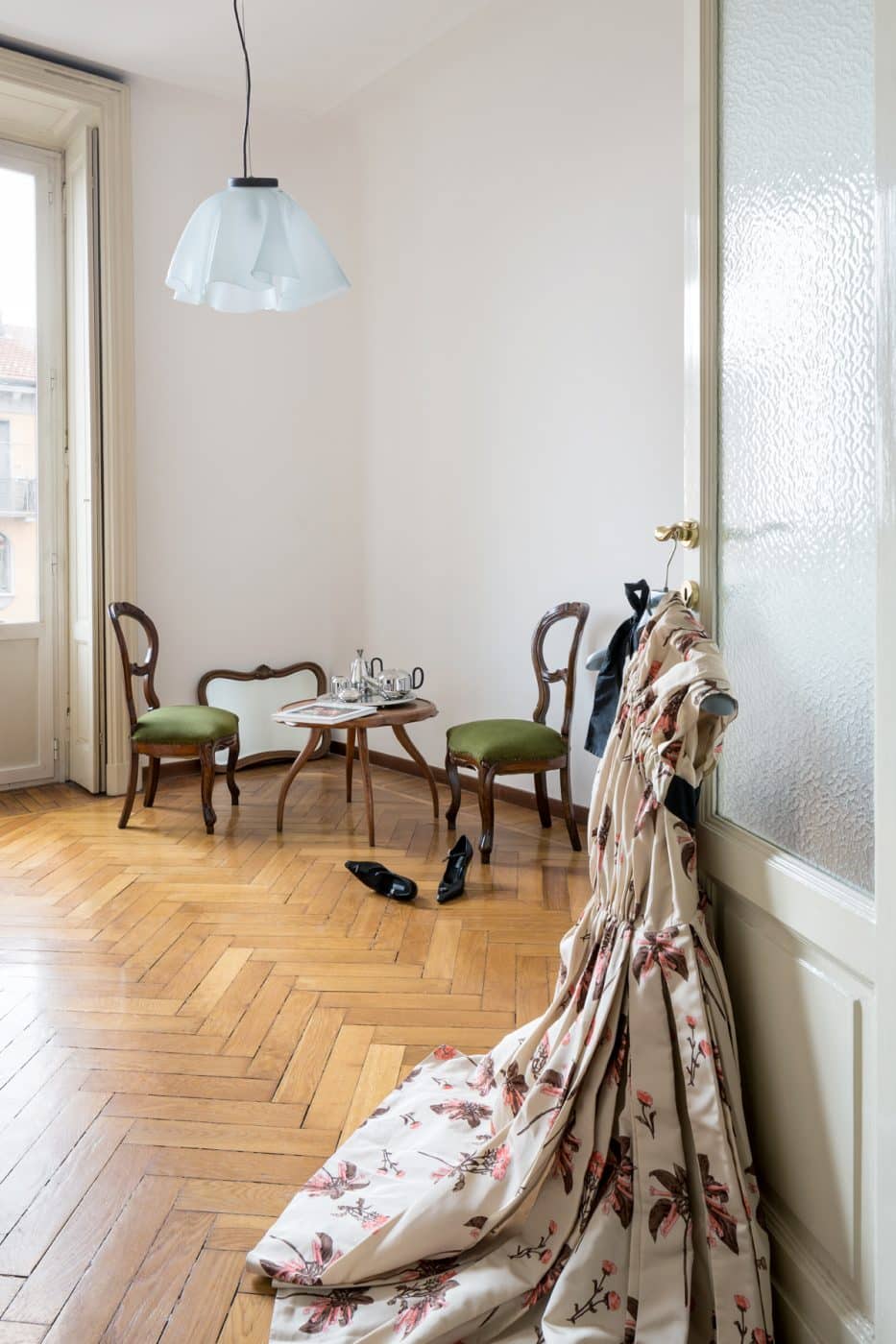 A view of Jenny Walton's Milan apartment with a cream-colored Prada dress with pink flowers hanging from a door handle, a 1960s wood-framed mirror against the wall, two baroque Italian chair with green upholstered seats and an Osvaldo Borsani cocktail table holding a silver-plated tray 