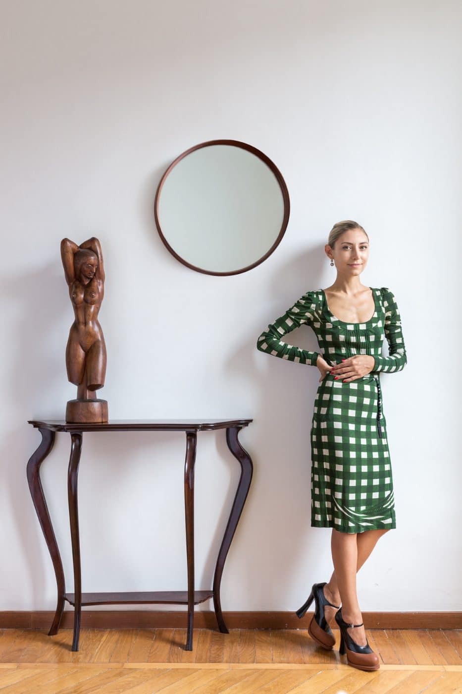 Walton in a green checked Prada dress standing next to a round wood-framed mirror hanging over an Italian mid-century console table topped by a wooden sculpture of a female nude