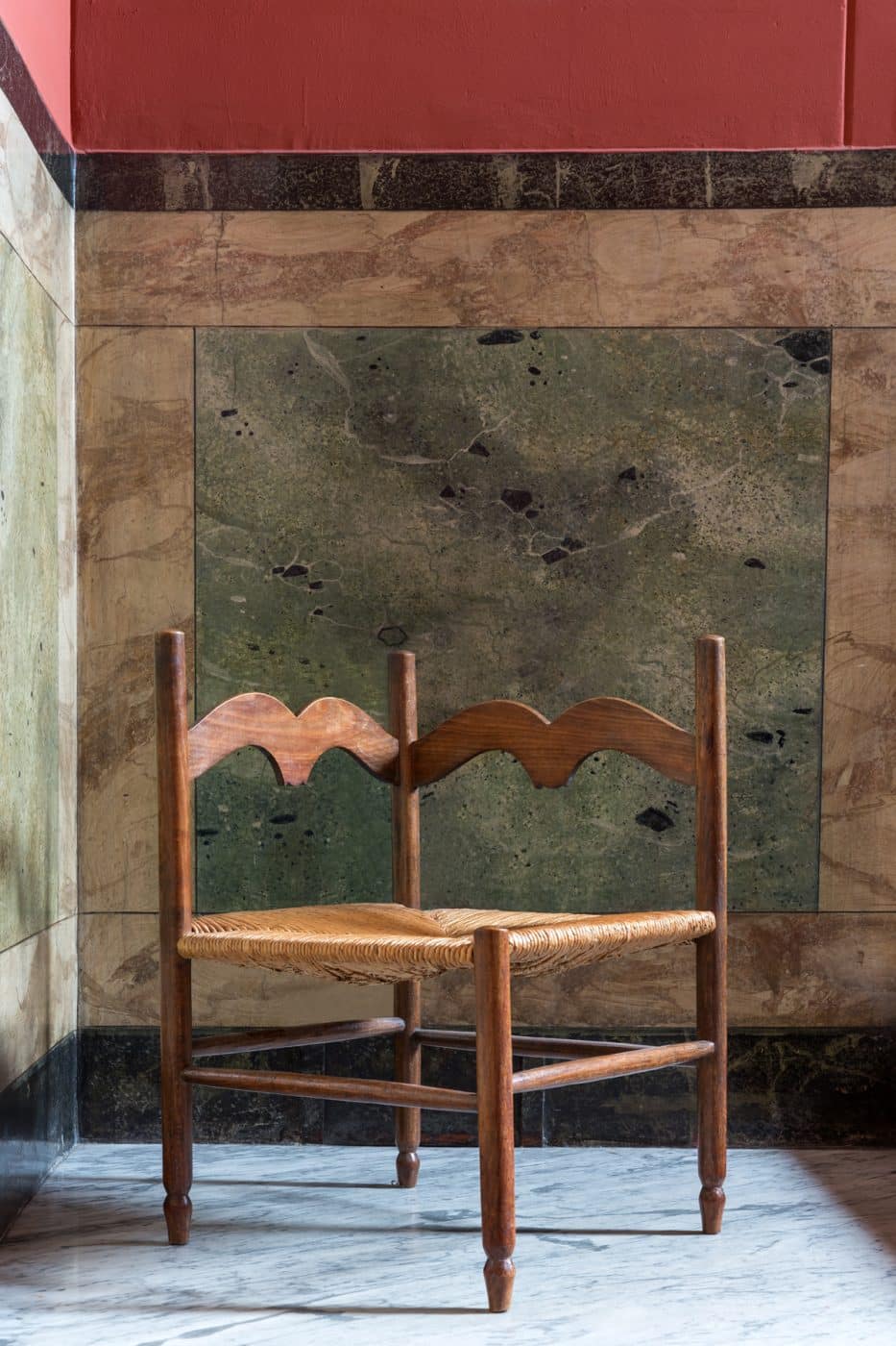 A French oak corner chair with a rush seat photographed against the colorful stone wall in the hallway of Jenny Walton's Milan apartment building
