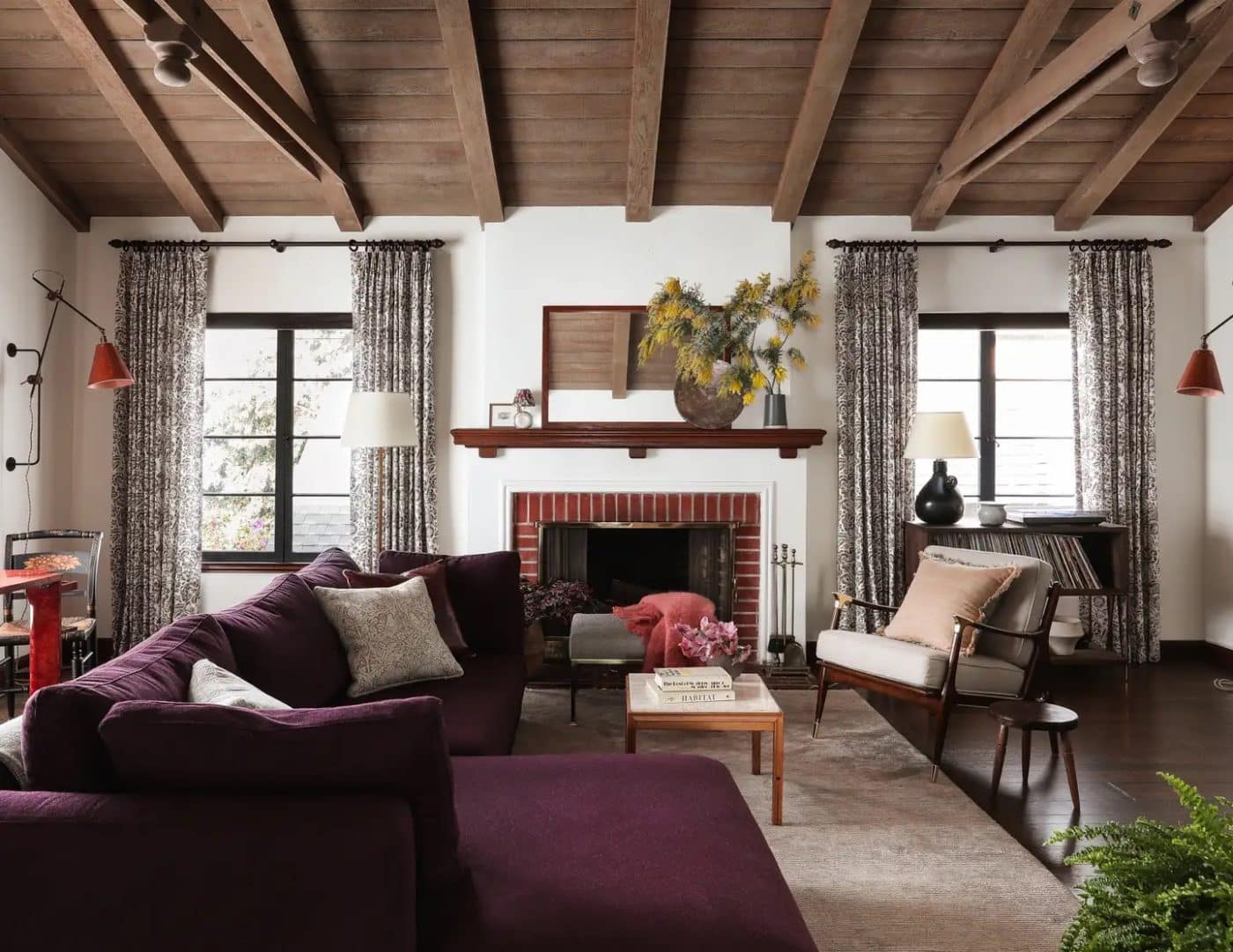 A living room with a wood-beam ceiling, off-white walls, a brick fireplace, a deep plum sofa and a wooden armchair and stool