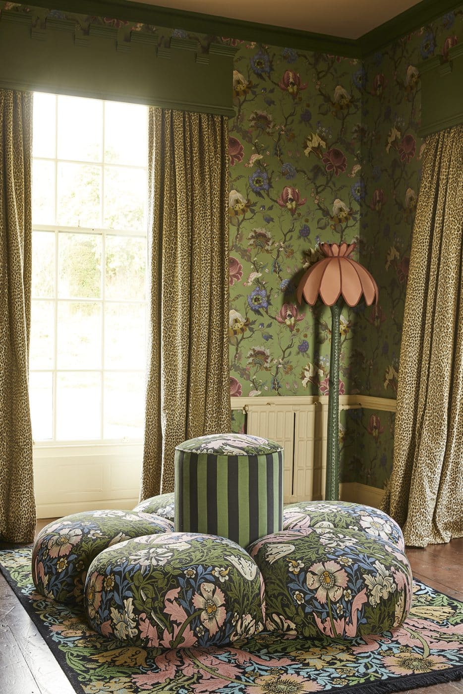 The drawing room at Trematon castle, decorated with green floral wallpaper, leopard-print curtains, the brand's Acanthus floor lamp, and a rug and round seating piece in a floral William Morris print
