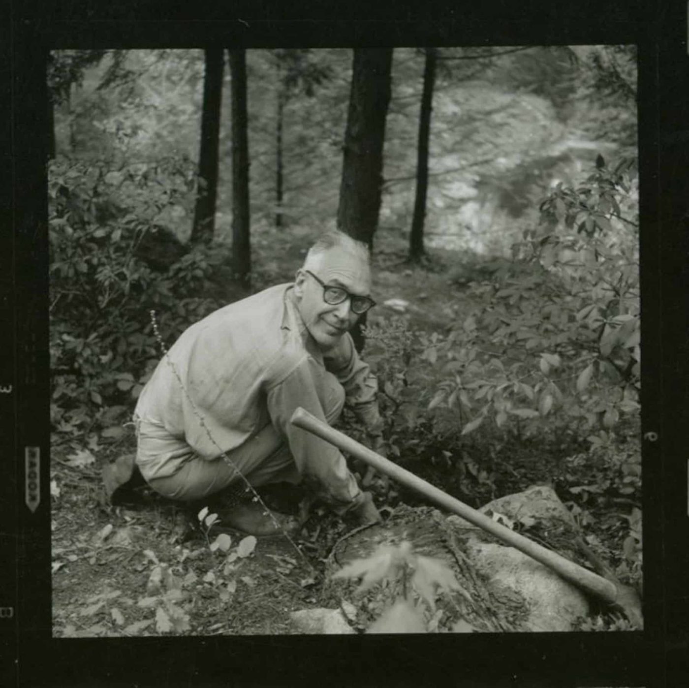 A black-and-white photo of Russel Wright at Manitoga, crouching outdoors, next to a pickax