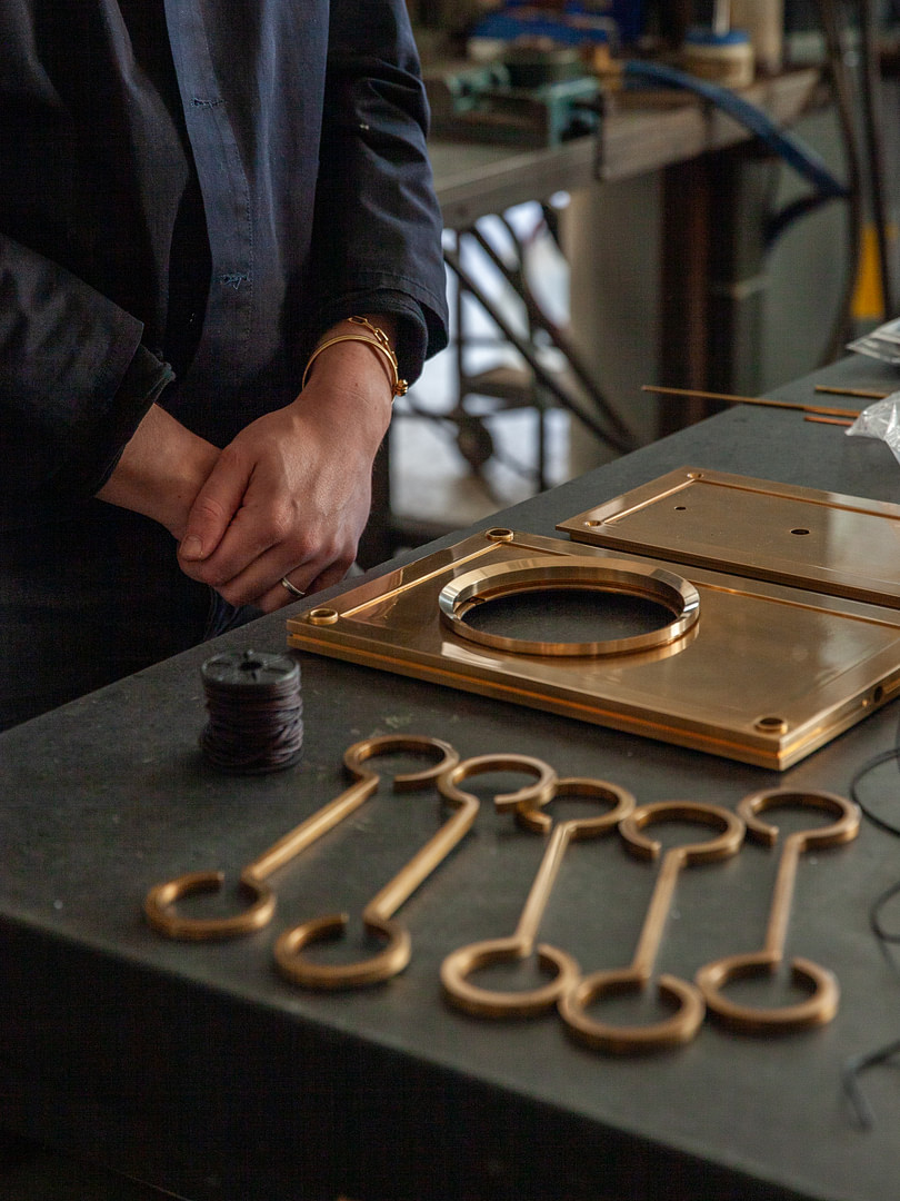 Fisher stands at a table bearing some of the elements used to make the lights in his Ariadne collection, namely a spool of leather cord and brass plates and hooks.