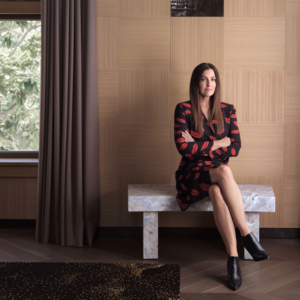 Interior designer Nicole Hollis wearing a black dress with a red lip print and sitting on a stone bench against a wood-paneled wall