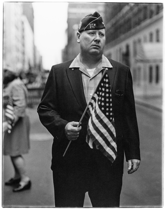 A black-and-white photo of an American veteran standing in the street holding a small American flag, by photographer Diane Arbus