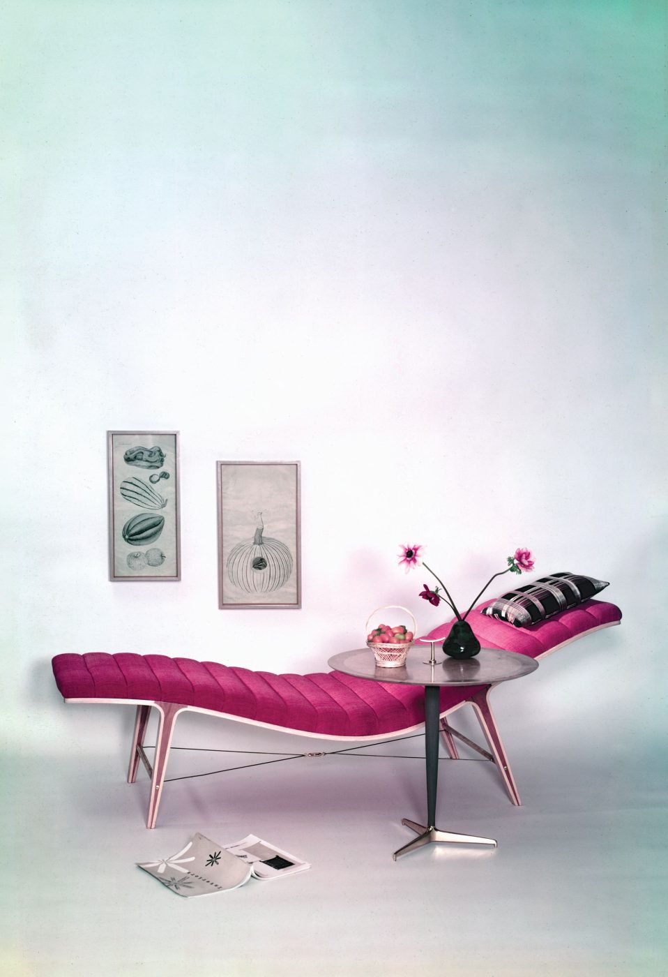 A vignette composed of a hot-pink chaise longue, a side table with a vase of flowers and a small decorative fruit basket, and a pair of monochromatic artworks on the wall