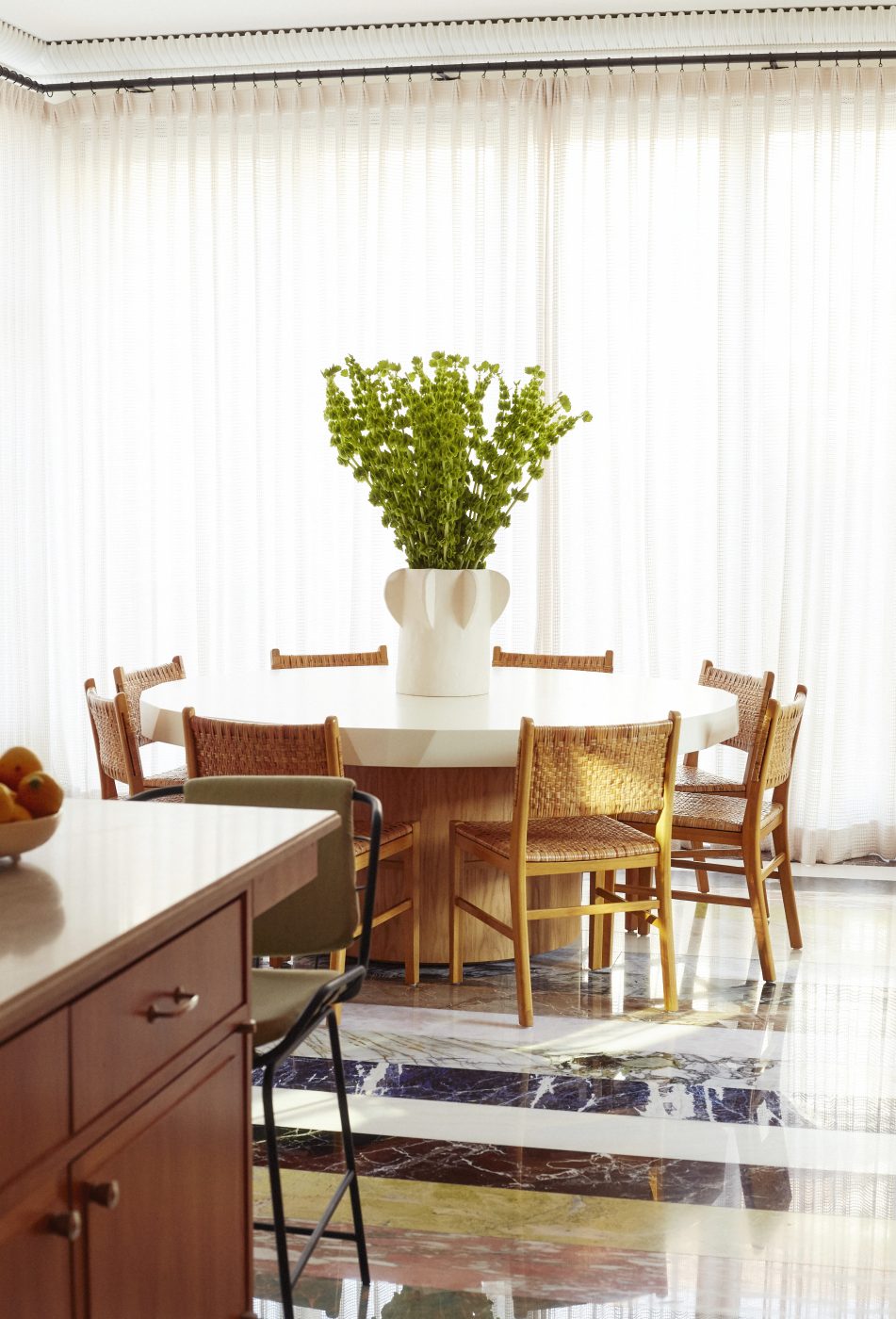 Woven-rattan chairs surround a barrel-based contemporary table in the dining area.of the kitchen of a home in Brooklyn's Gravesend neighborhood with interior design by Eli Dweck 