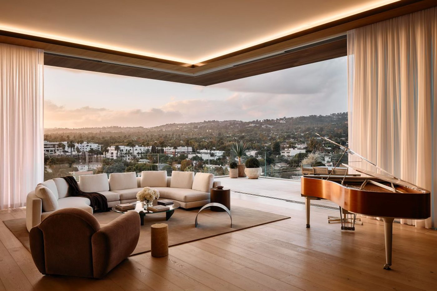 Corner living area in penthouse in Los Angeles West Hollywood designed by Adair Lewis of JSN Studio with corner walls of glass that retract to open to balcony. Furnished with grand piano, curving white couch, metal coffee tables.