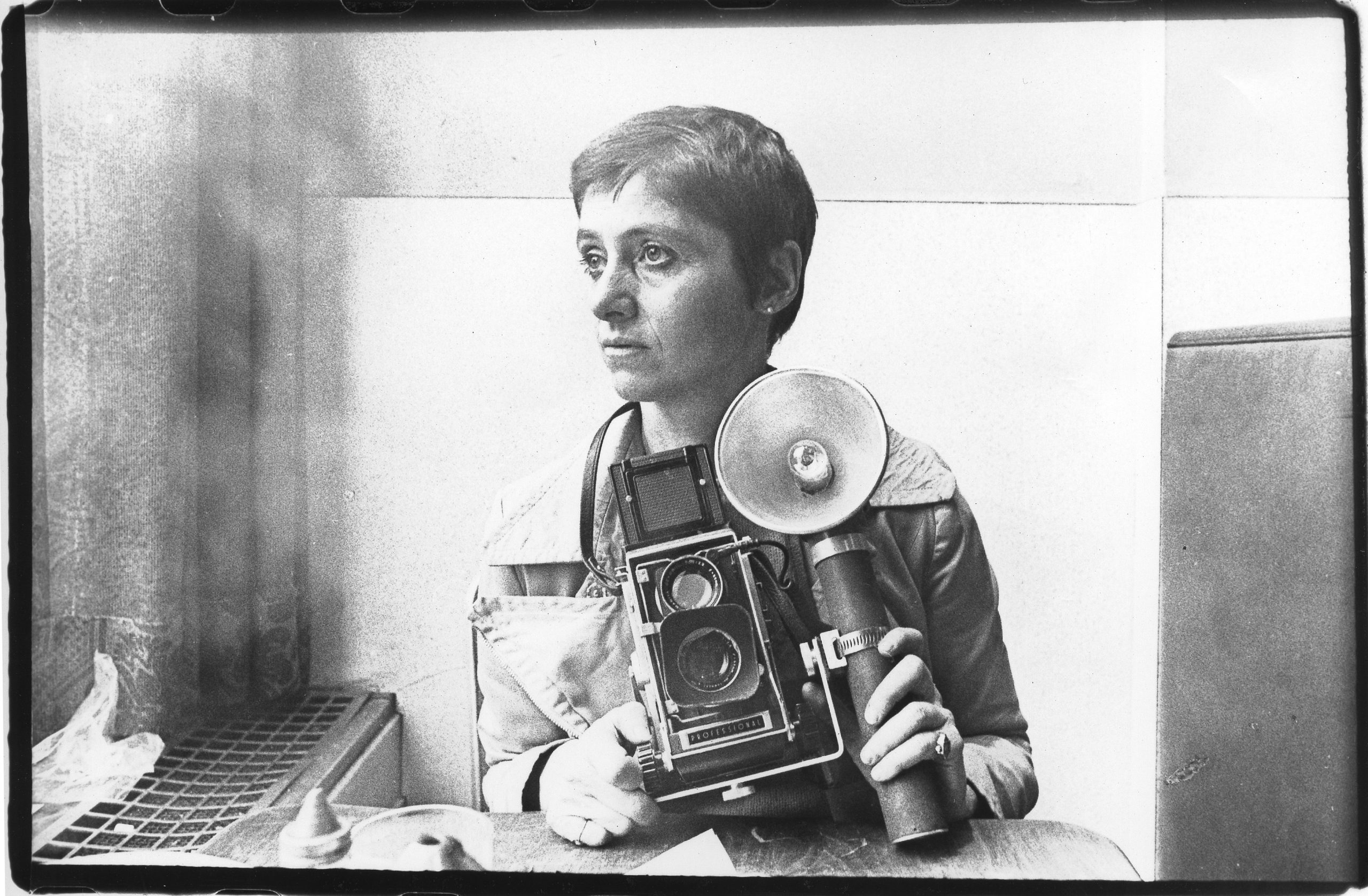 A black-and-white photo of photographer Diane Arbus sitting at a restaurant table and holding her camera