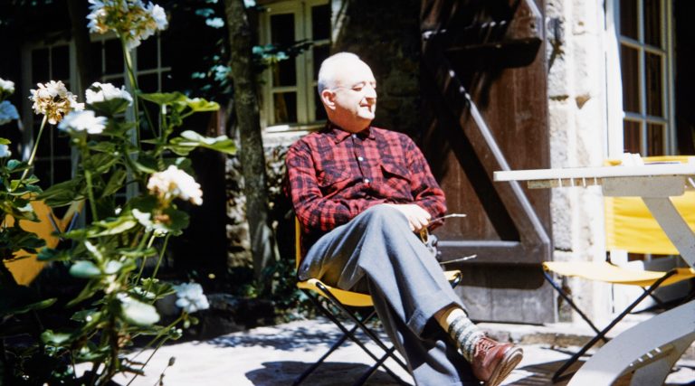 Jean Royère wearing a red plaid shirt and sitting in a yellow folding chair in the garden of his home in Brittany