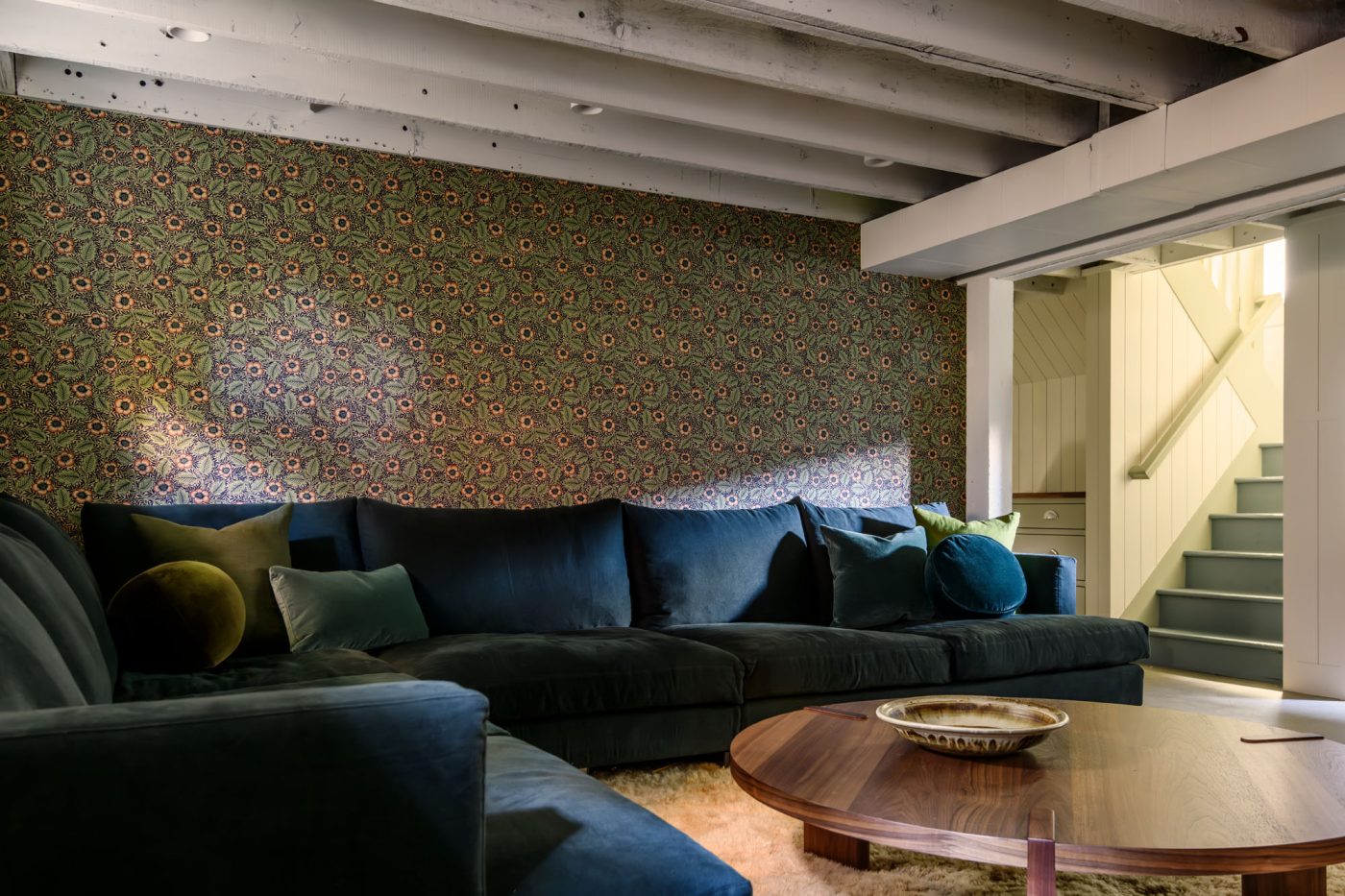 Basement with large blue corner sectional sofa, low wooden circular coffee table, white shag rug and floral wallpaper in a house in Seattle's Capitol Hill neighborhood designed by Lisa Staton