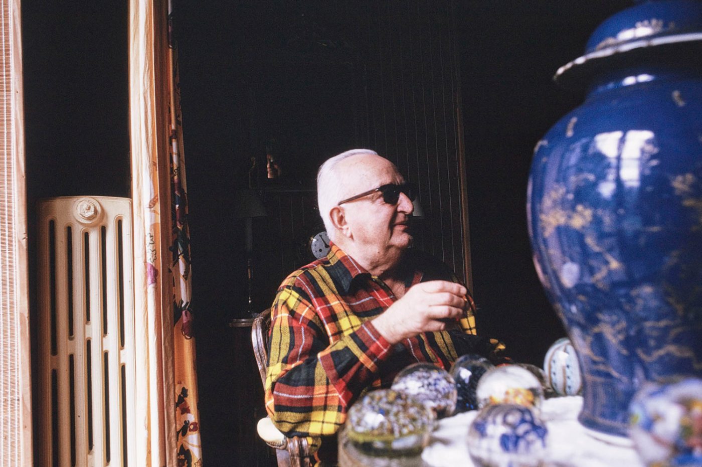 Jean Royère wearing sunglasses and a red-and-yellow plaid shirt sitting at a table near a window in his home in Brittany