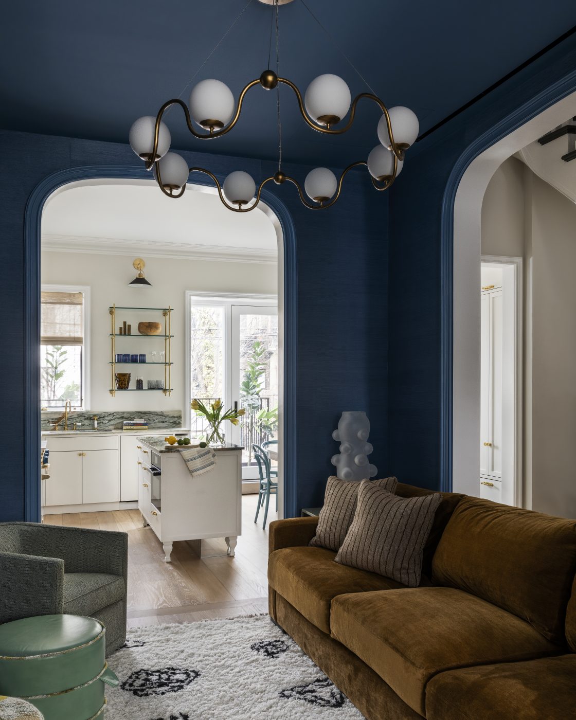 The cozy living room has deep blue walls and built-in shelving, a pair of A. Rudin barrel-back chairs, two Nickey Kehoe poufs, a tattoo-patterned Nordic Knots rug, a wavy glass-globed Currey and Company chandelier and a Kravet sofa.