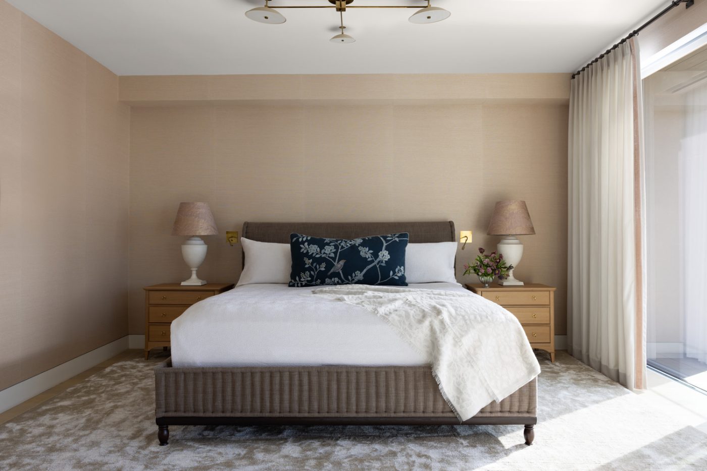 Primary bedroom of a San Francisco row house designed by Marea Clark featuring a spare style, to keep the focus on the views through the floor-to-ceiling windows. Oak nightstands from Nickey Kehoe, topped by handmade ceramic lamps, flank an oak bed covered in Belgian linen. The hemp grasscloth wallcovering, by Phillip Jeffries, creates a sense of cocooned serenity.
