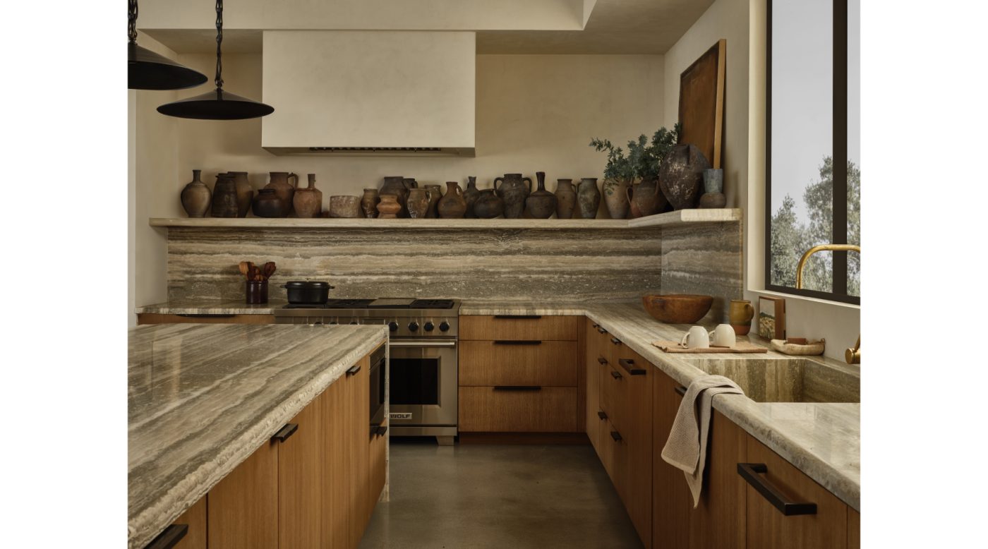 Kitchen of Malibu California home of NFL player Baltimore Ravens Ronnie Stanley designed by Sarah Solis featuring travertine counters and backsplash and collection of clay pots and vessels on shelf over the range