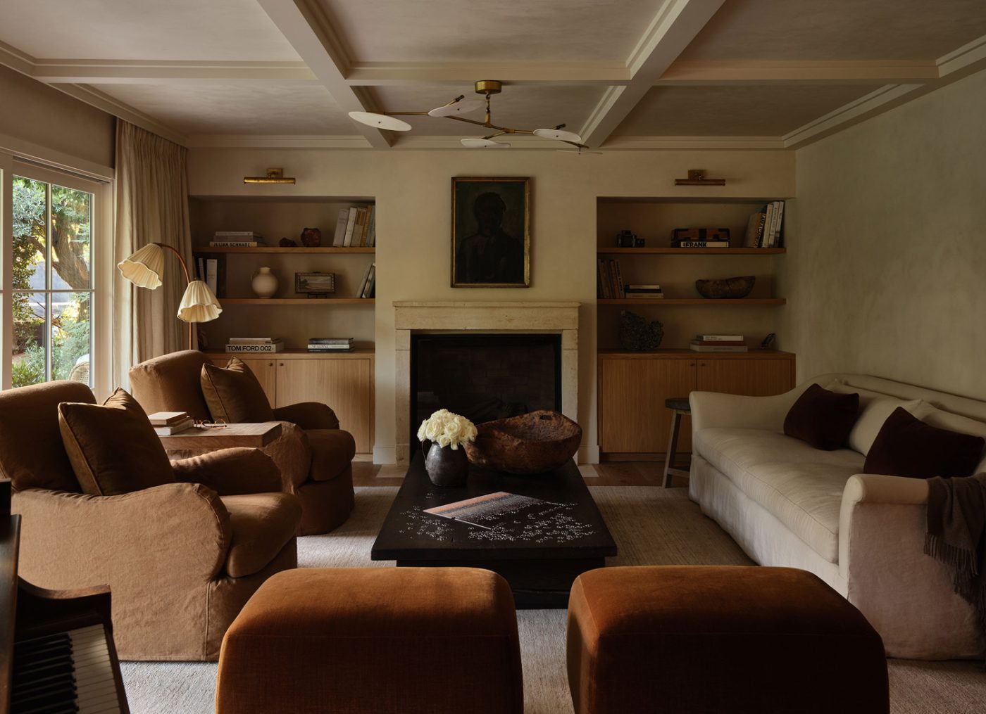 Earth-toned living room of Malibu California home of interior designer Sarah Solis featuring pieces from her Galerie Solis: a Classic sofa, a Relic coffee table, Royal lounge chairs, in front of an antique limestone fireplace mantel from Pittet Architecturals. The chandelier is by Lindsey Adelman, and the 1940s Swedish floor lamp is from 1stDibs.