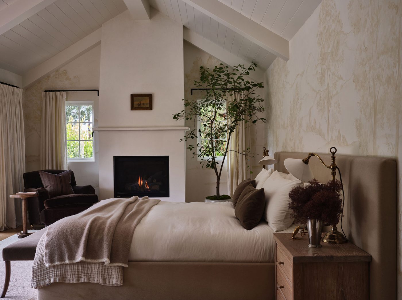 Primary bedroom of Malibu California home of interior designer Sarah Solis featuring lamps from Jamb, a mohair-upholstered Galerie Solis bed, a George Smith lounge chair in a corner. The mural behind the bed was hand-painted by artist James Mobley.