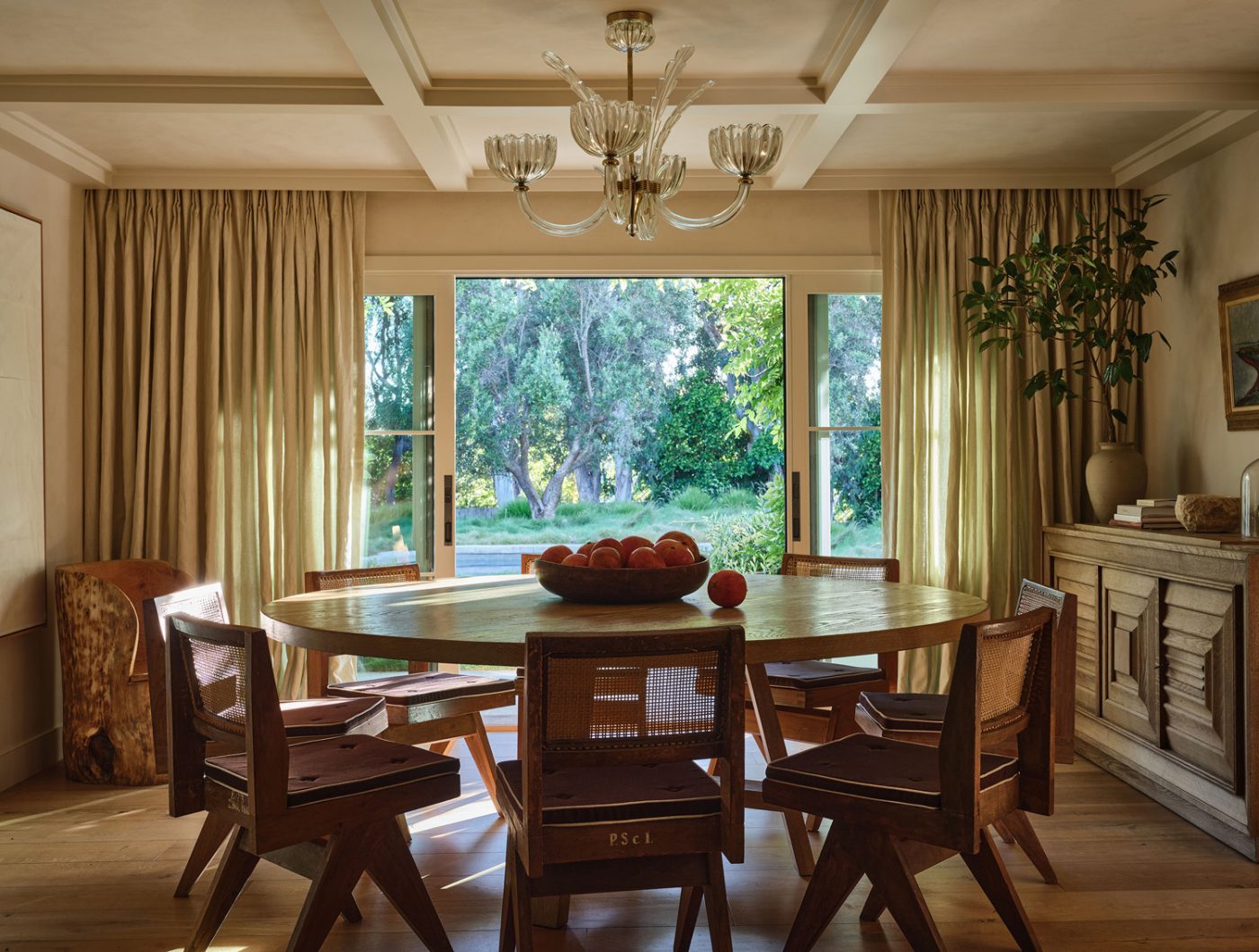 Earth-toned dining room of Malibu California home of interior designer Sarah Solis featuring Pierre Jeanneret chairs around a Galerie Solis Story table beside a 1940s Charles Dudouyt sideboard. The chandelier is Murano glass from the 1960s, the drapery fabric is Pierre Frey, and the painting above the sideboard is a vintage Swedish work.