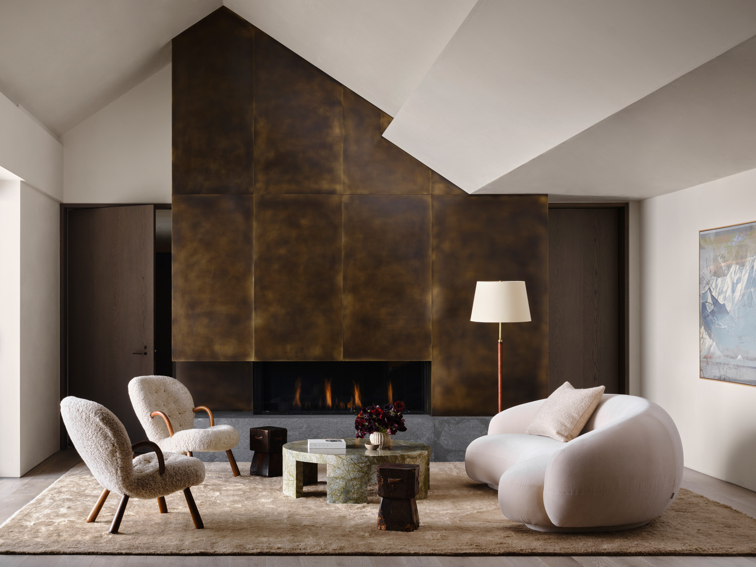 primary suite sitting area of a house designed by Vanessa Alexander in Aspen, Colorado, featuring bronze-colored paneled fireplace surround behind a curving white TacChini sofa, green marble coffee table  by Francesco Balzano and shearling covered Clam chairs by Arnold Madsen