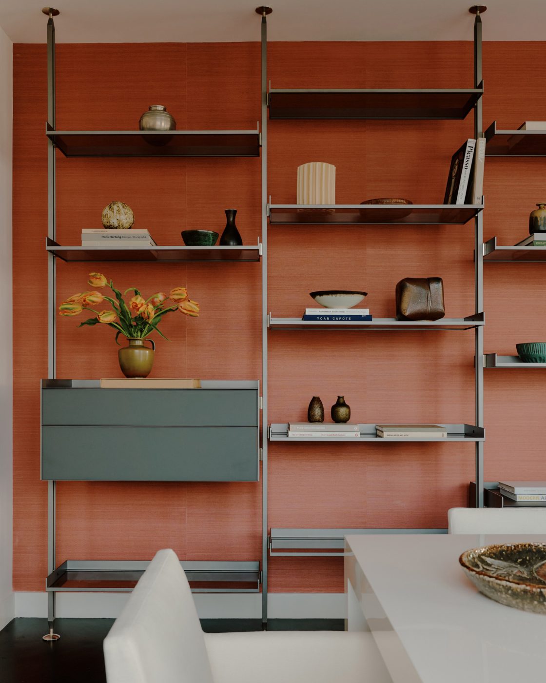 Dining room in nearly all white minimalist loft apartment designed by Clive Lonstein and featuring a Rimadesio tension-mounted floor-to-ceiling shelving unit against a salmon-colored accent wall.