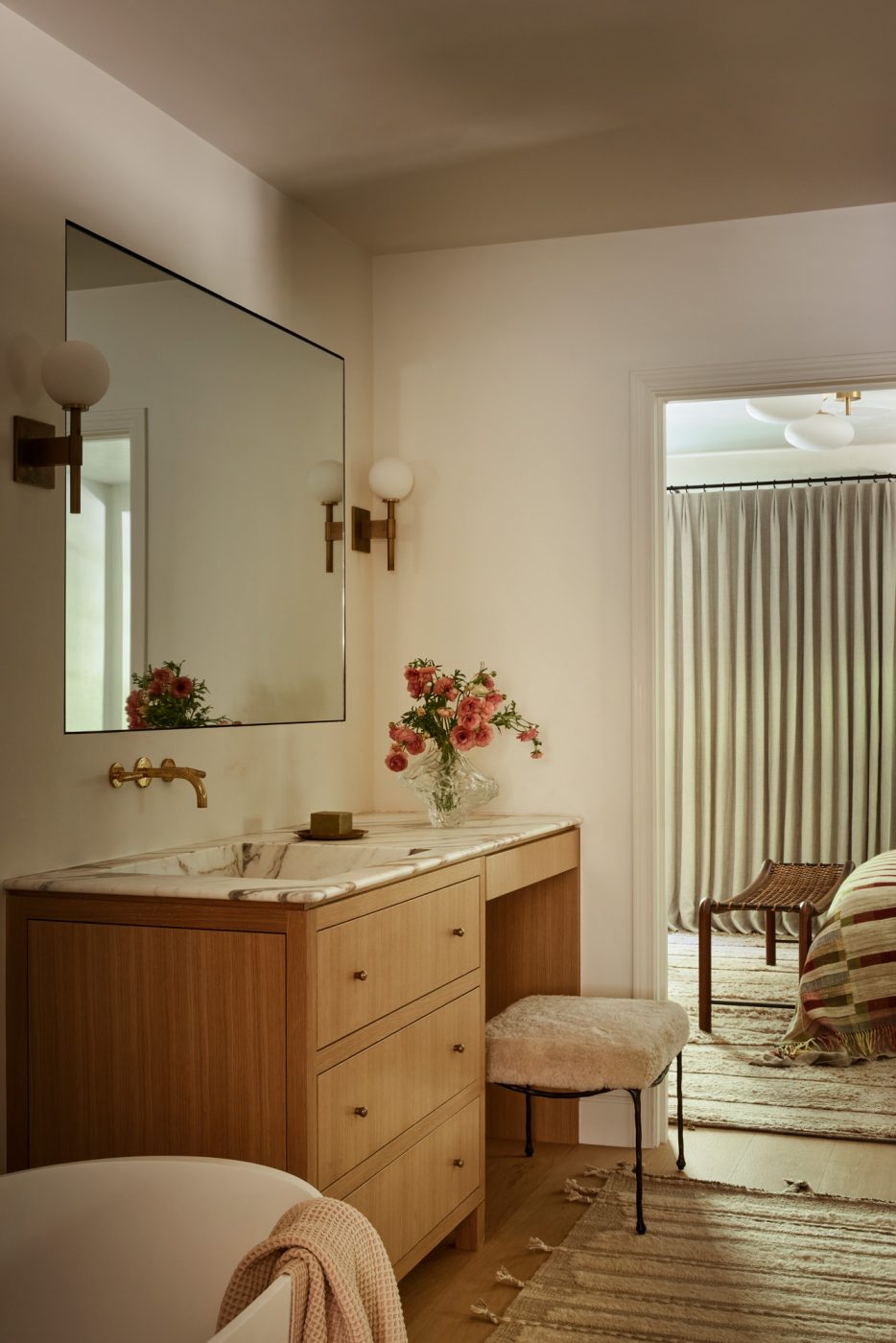 Primary bathroom in Beverly Hills home designed by Maggie Smith with layered textures, warm woods, a marble-topped vanity, Allied Maker sconces and a custom mirror.