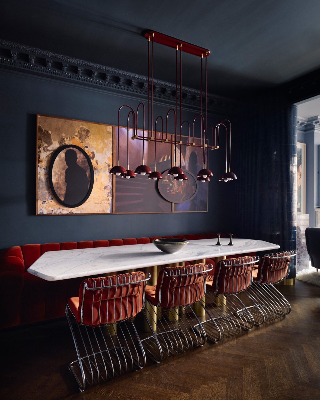 Goat fur rug, orange postmodern sofa, contemporary steel coffee Marble topped table and red upholstered banquet plus bent steel chairs and chandlier in Dark blue walled dining room in Harlem townhouse designed by Crystal Sinclair
