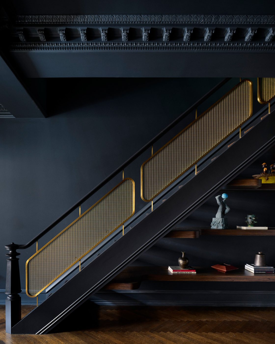 Staircase with new modern-looking brass railing and shelves built in underneath displaying sculptures in dining room in Harlem townhouse designed by Crystal Sinclair