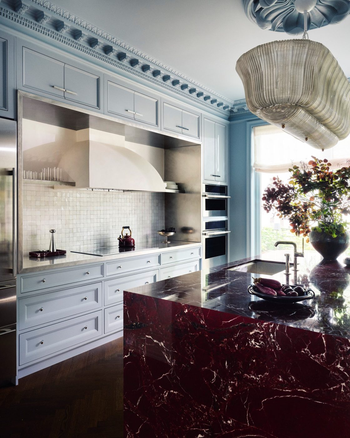 Polished red marble island in pale blue-gray kitchen with elaborate moldings, Murano chandelier and herringbone floors in a Harlem townhouse design by Crystal Sinclair