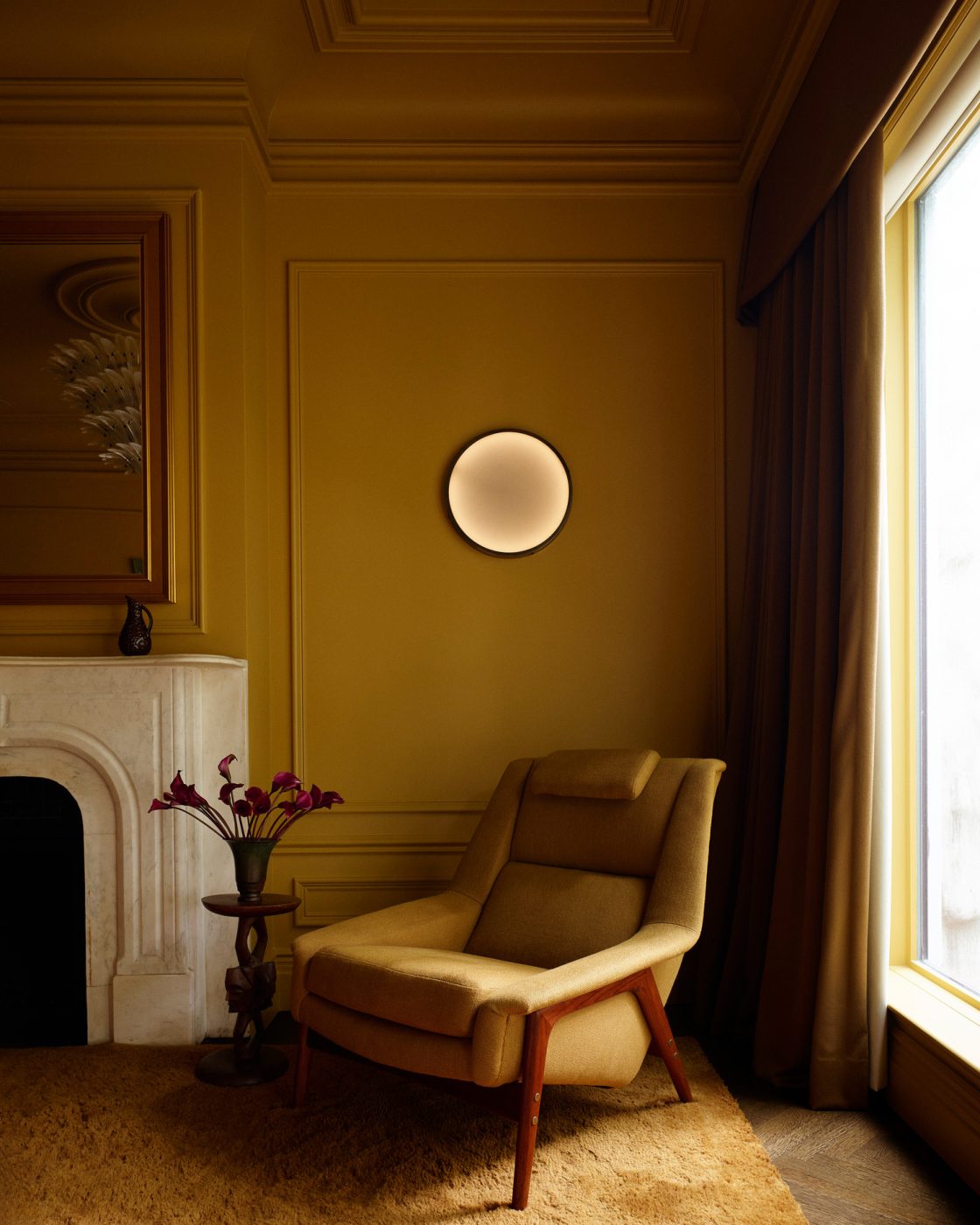 Seating area with Folke Ohlsson for Dux lounge chair beside white marble fireplace in deep yellow primary bedroom in a Harlem townhouse design by Crystal Sinclair