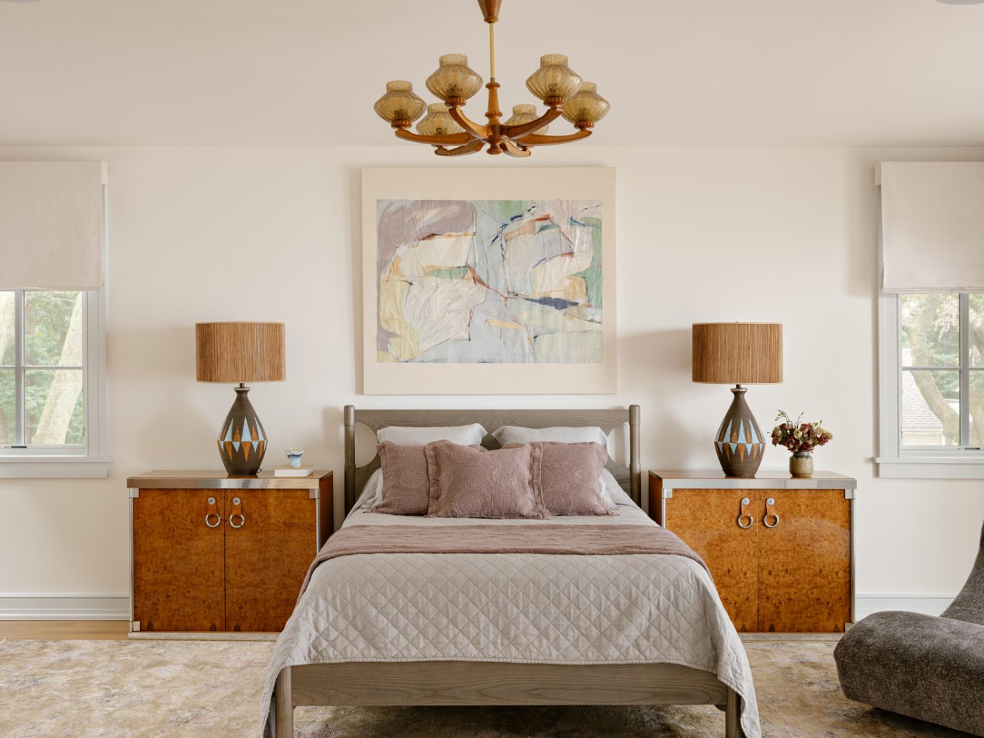 1960 Bitossi ceramic lamps, a pair of 1970s Guido Faleschini burl-wood sideboards flanking the bed, artwork by Anne Wehrley Björk, and a Danish carved-oak six-light chandelier with amber art-glass shades in the primary bedroom of a Long Island home with interiors by studio Evan Edward designers Michael Edward Moirano and Josh Evan Goldfarb