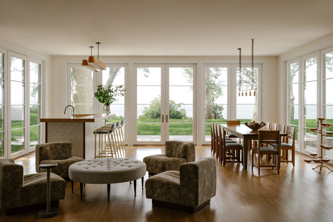 A lounging area with a mid-20th-century button-tufted ottoman and a set of four of low 1970s chairs by Tito Agnoli in the window-walled sunroom bar of a Long Island home with interiors by studio Evan Edward designers Michael Edward Moirano and Josh Evan Goldfarb