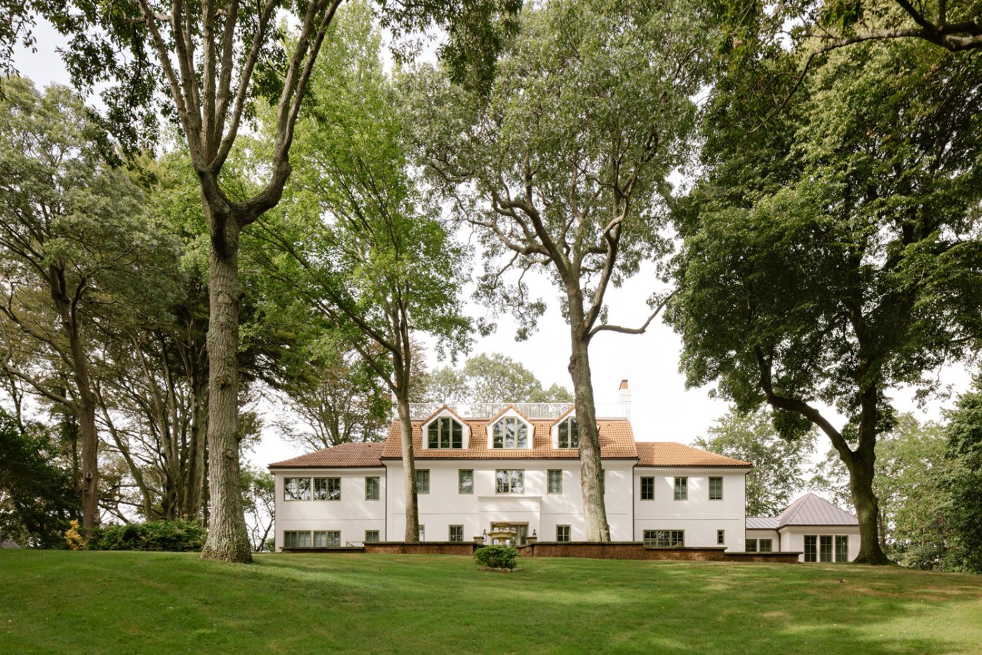 Rear exterior and tree-dotted lawn of a mediterranean-style historic home on Long Island's North Shore with interior redone by studio Evan Edward interior designers Michael Edward Moirano and Josh Evan Goldfarb