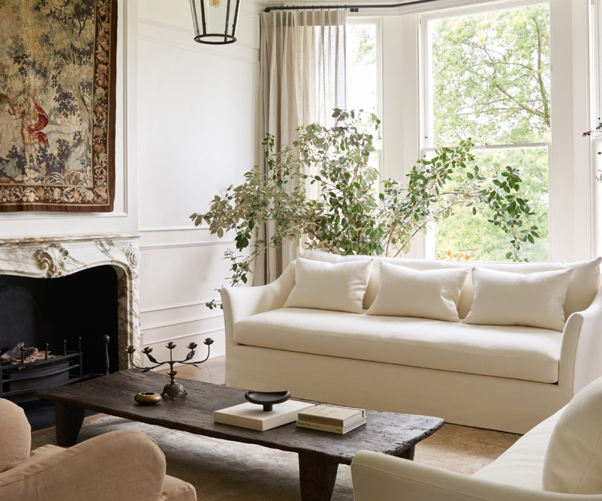 Sun-filled nearly all white living room with a pair of bespoke ivory linen sofas flanking an antique farm table and a 19th-century Aubusson tapestry above a Breccia marble fireplace, in an Italianate house in London’s Notting Hill with interior design by Katie Harbison.