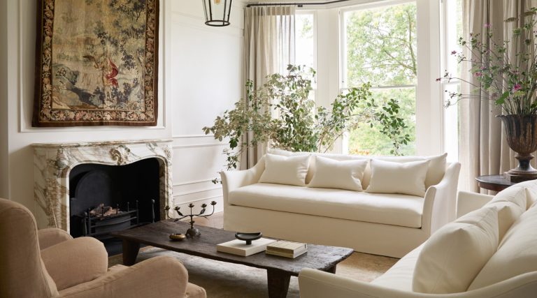 Sun-filled nearly all white living room with a pair of bespoke ivory linen sofas flanking an antique farm table and a 19th-century Aubusson tapestry above a Breccia marble fireplace, in an Italianate house in London’s Notting Hill with interior design by Katie Harbison.