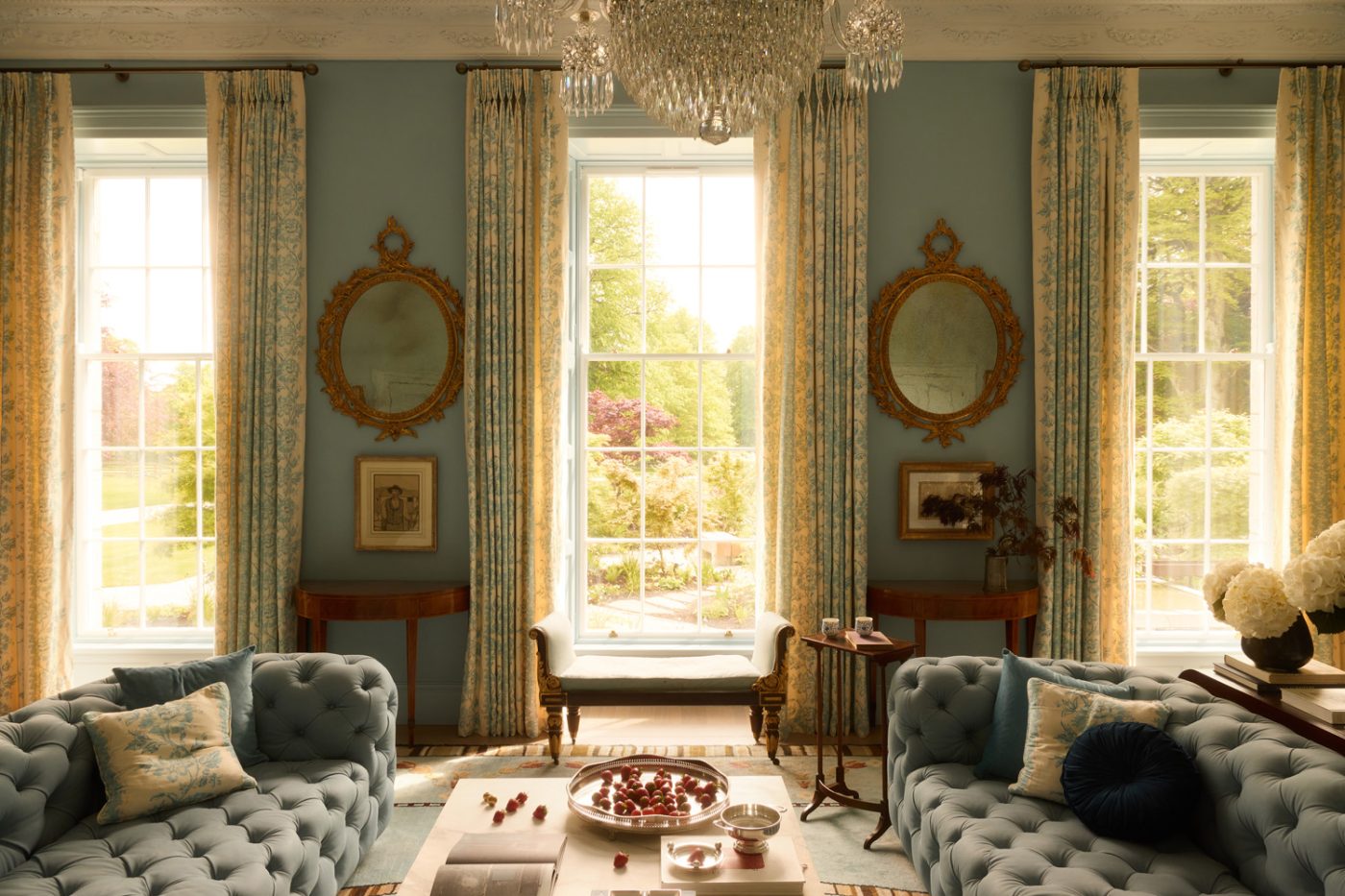 Contemproary chesterfield couches and tall windows under a Regency crystal chandelier in blue drawing room of 19th-century Georgian-style manor house in Irish Countryside designed by Dublin-based Róisín Lafferty
