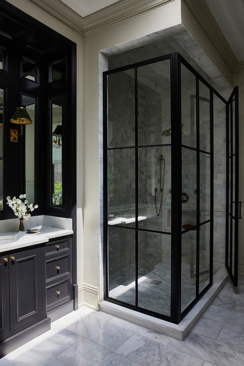 A dark vanity with white counter, light gray marble floors and walls, and a metal-and-glass stall shower enclosure in the primary bathroom of a historic Chicago townhouse by design partners Laura Buxbaum Gordon and Nate Berkus
