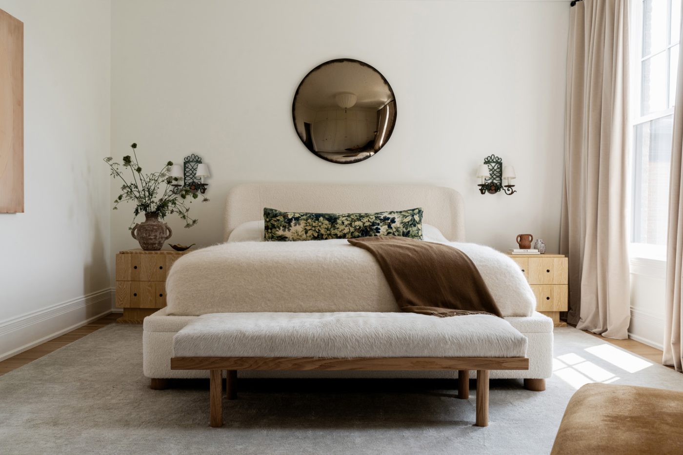 A convex bronze Marmorea London mirror hangs on the wall behind a custom upholstered bed flanked by Lawson-Fenning nightstands and barley-twist sconces in the primary bedroom of a historic Chicago townhouse by design partners Laura Buxbaum Gordon and Nate Berkus