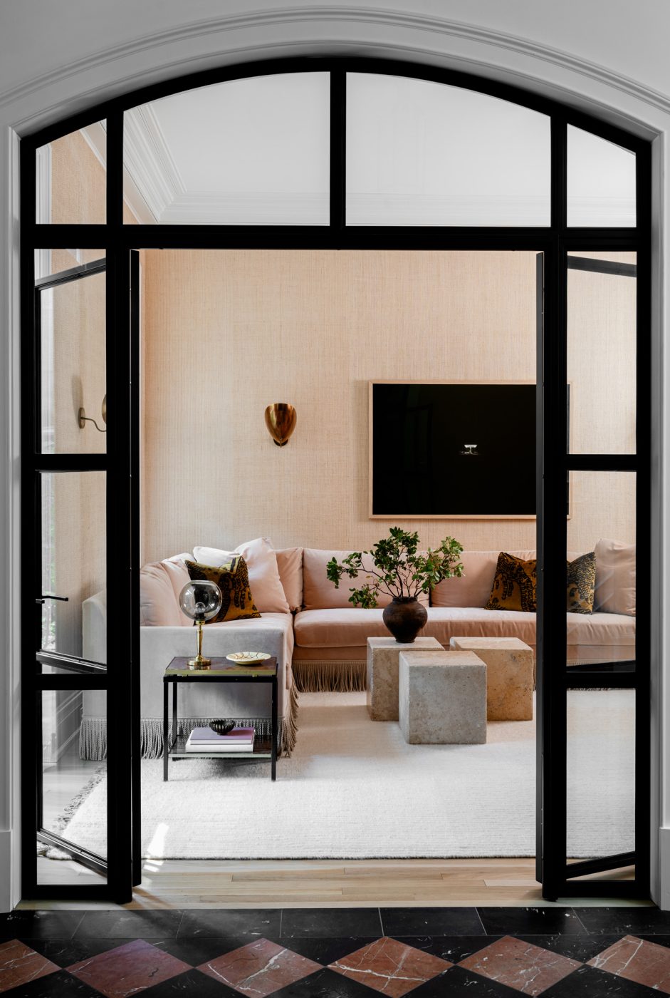 Fringed sectional sofa and other neutral furniture seen through iron-and-glass partially opened doors in the sitting room/wine lounge of a historic Chicago townhouse by design partners Laura Buxbaum Gordon and Nate Berkus